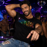 A man in a nightclub wearing a black t-shirt with 'SHOWGURL' written across the chest in rainbow rhinestones by Rico Starr