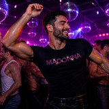 Man in a black t-shirt with 'MADONNA' in pink rhinestone text at a disco party by Rico Starr