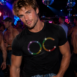 A man in a crowded nightclub wearing a black t-shirt with a rainbow-colored 'OC' graphic on the chest, surrounded by shirtless men and disco balls under blue and purple lighting by Rico Starr