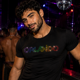 A man in a nightclub wearing a fitted black t-shirt with 'ORLANDO' printed across the chest in rainbow rhinestones by Rico Starr