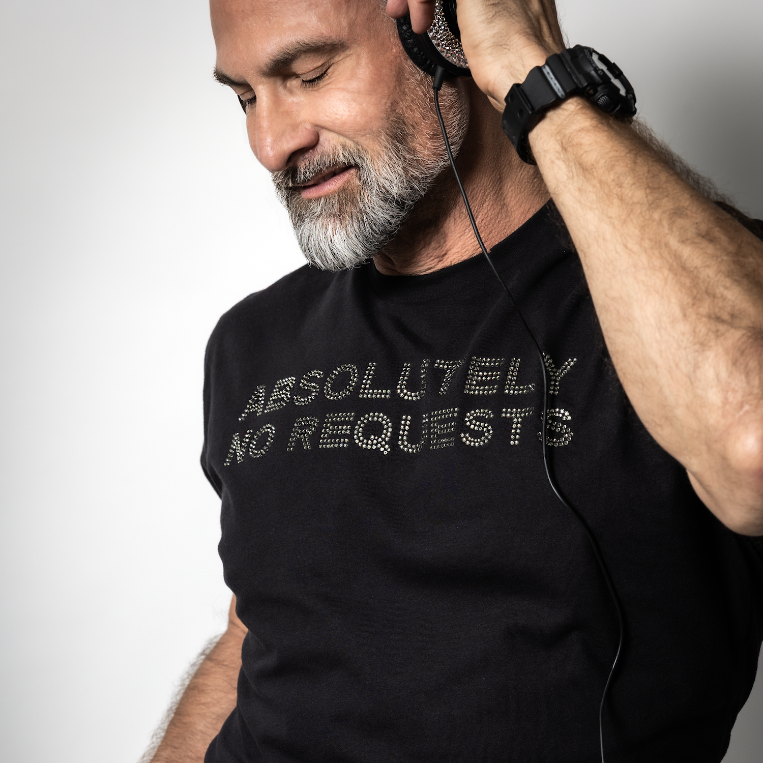 Male DJ wearing a black rhinestone “Absolutely No Requests” t-shirt while listening to music through headphones by Rico Starr