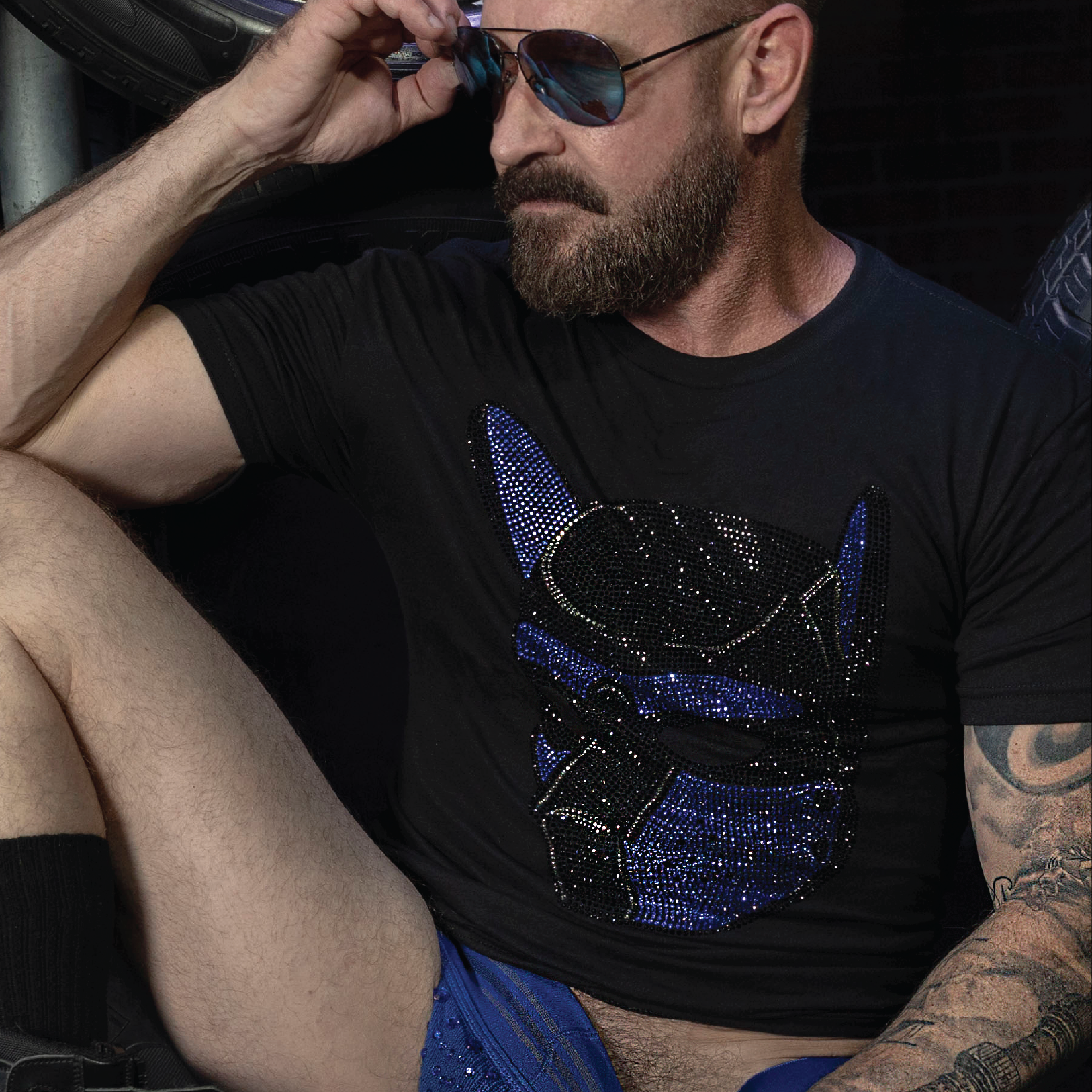 Man wearing a black t-shirt featuring a blue and black rhinestone pup mask graphic. LGBTQ+ fetish fashion inspired by pup play, leather culture, and gay nightlife, with sparkling crystal details, a fitted modern cut, and bold clubwear styling by Rico Starr