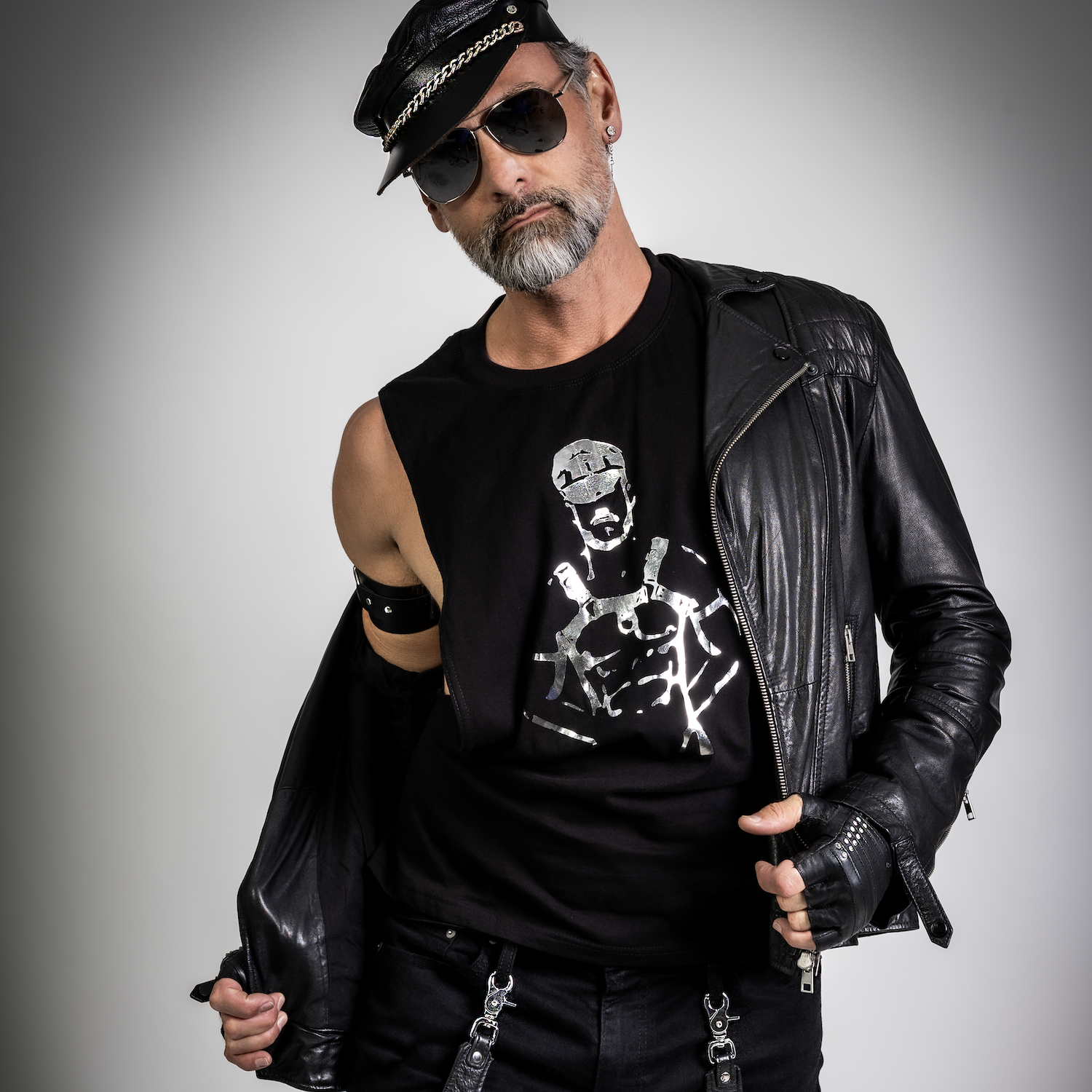 Male model wearing a black leather-inspired shredder tank top with a metallic fetish graphic, styled with a biker cap, leather jacket, gloves, and sunglasses in an edgy gay club fashion look by Rico Starr