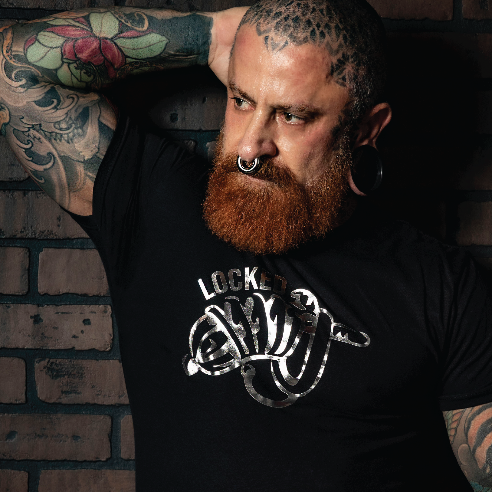 Bearded tattooed man wearing a black "Locked" graphic t-shirt with metallic silver fetish-inspired artwork. Gay leather and kink fashion featuring a bold chastity-themed design, fitted modern cut, and LGBTQ+ nightlife style for fetish events, Pride celebrations, and alternative men's fashion by Rico Starr