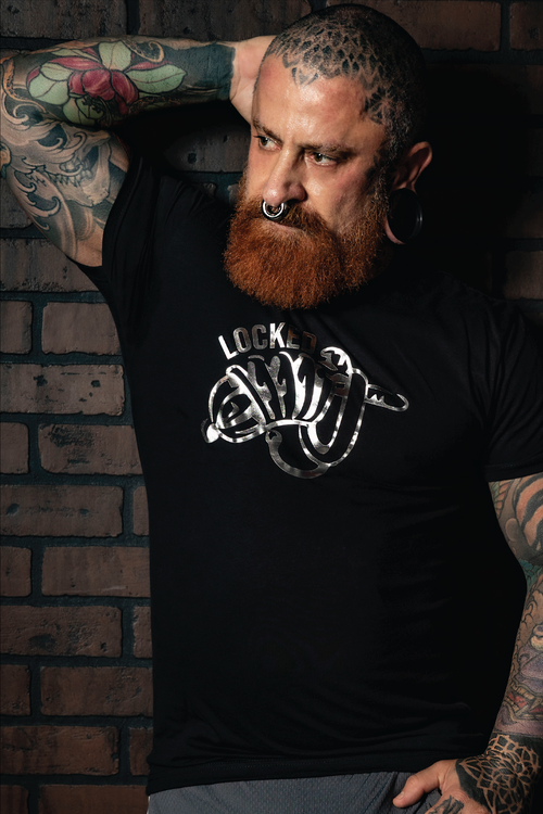 Bearded tattooed man wearing a black "Locked" graphic t-shirt with metallic silver fetish-inspired artwork. Gay leather and kink fashion featuring a bold chastity-themed design, fitted modern cut, and LGBTQ+ nightlife style for fetish events, Pride celebrations, and alternative men's fashion by Rico Starr