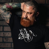 Bearded tattooed man wearing a black "Locked" graphic t-shirt with metallic silver fetish-inspired artwork. Gay leather and kink fashion featuring a bold chastity-themed design, fitted modern cut, and LGBTQ+ nightlife style for fetish events, Pride celebrations, and alternative men's fashion by Rico Starr