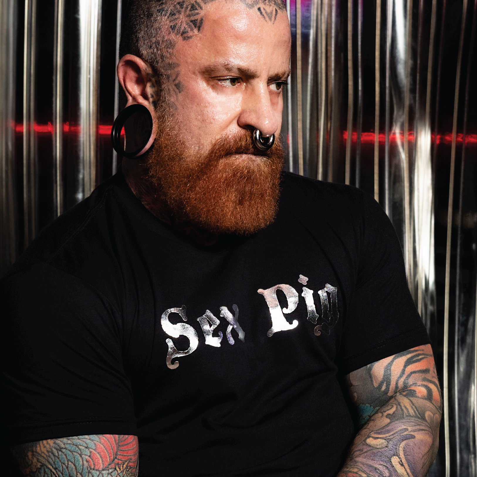 Tattooed bearded man wearing a black "Sex Pig" graphic t-shirt with metallic silver lettering. Bold LGBTQ+ fetish fashion inspired by leather, bear, and kink culture, featuring a fitted modern cut and edgy nightlife style for gay men, Pride events, leather weekends, and alternative fashion scenes by Rico Starr