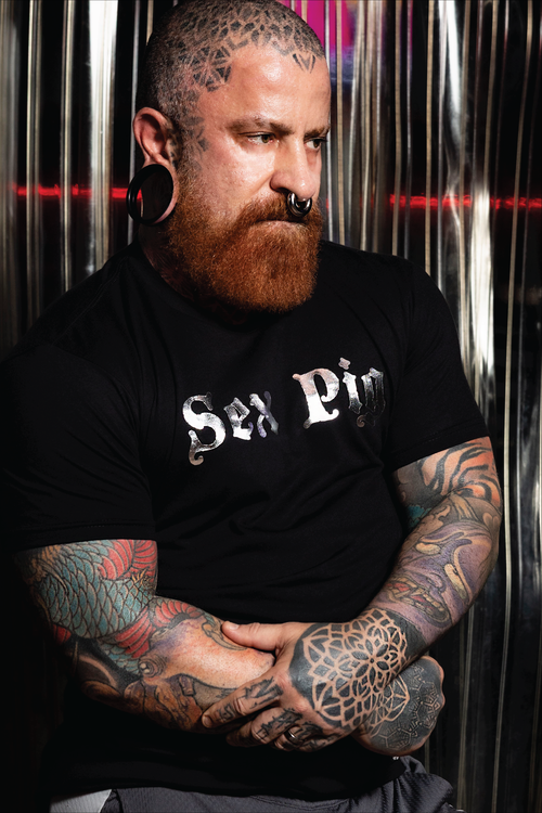 Tattooed bearded man wearing a black "Sex Pig" graphic t-shirt with metallic silver lettering. Bold LGBTQ+ fetish fashion inspired by leather, bear, and kink culture, featuring a fitted modern cut and edgy nightlife style for gay men, Pride events, leather weekends, and alternative fashion scenes by Rico Starr