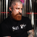 Tattooed bearded man wearing a black "Sex Pig" graphic t-shirt with metallic silver lettering. Bold LGBTQ+ fetish fashion inspired by leather, bear, and kink culture, featuring a fitted modern cut and edgy nightlife style for gay men, Pride events, leather weekends, and alternative fashion scenes by Rico Starr
