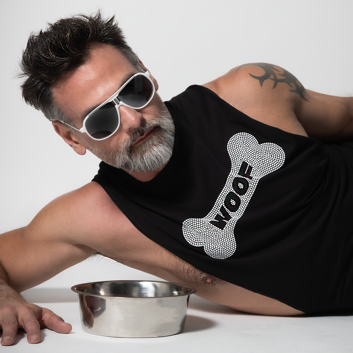 Silver fox male model wearing a black cut-off tank top with rhinestone “WOOF” dog bone graphic and white sunglasses beside a metal dog bowl by Rico Starr